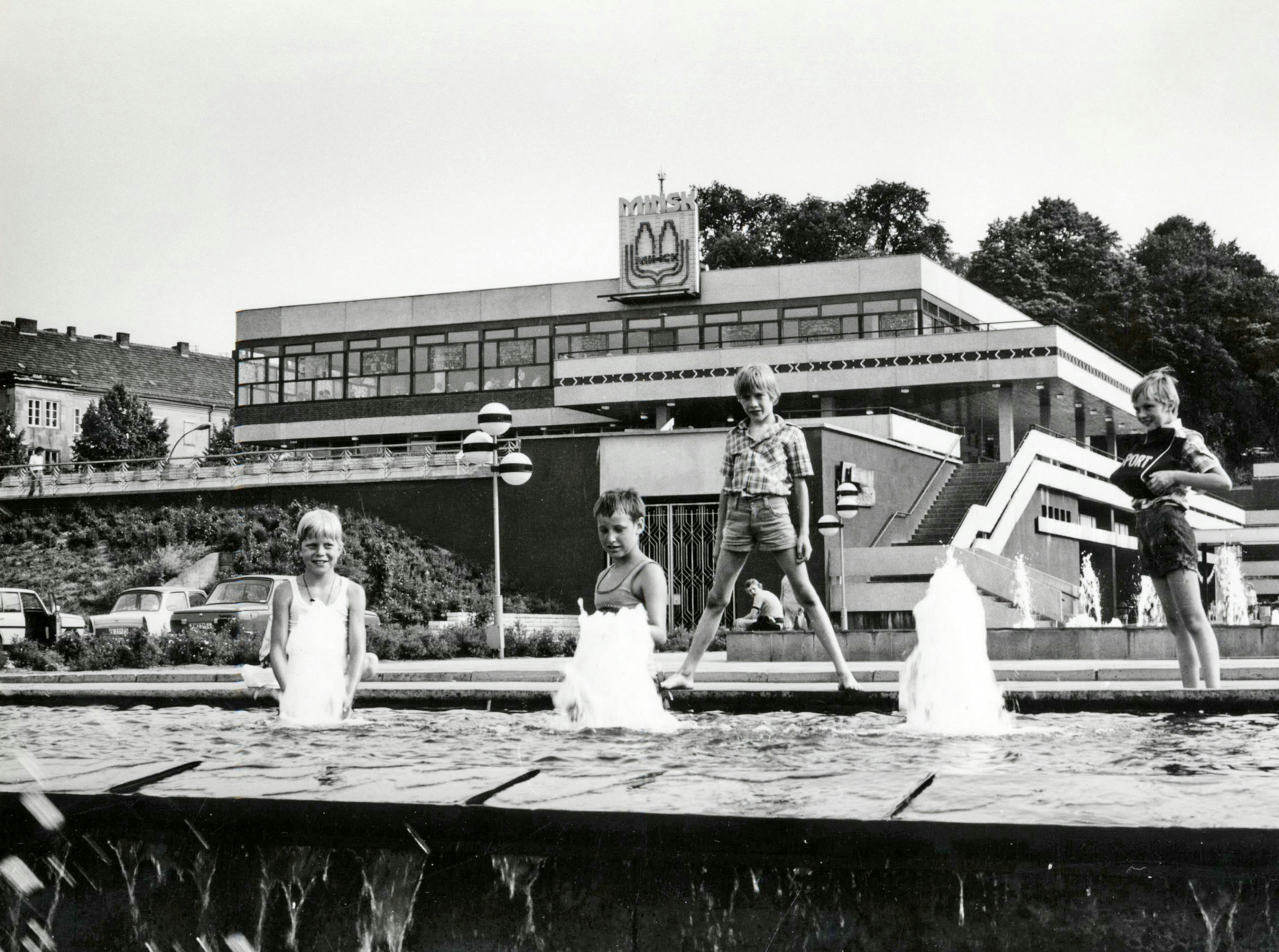 Schwarz-weiß-Foto: im Vordergrund spielen vier sommerlich gekleidete Kinder an einem Springbrunnen. Ein Junge steht breitbeinig auf seinem Rand. Im Hintergrund das ehemalige Terrassenrestaurant Minsk. 